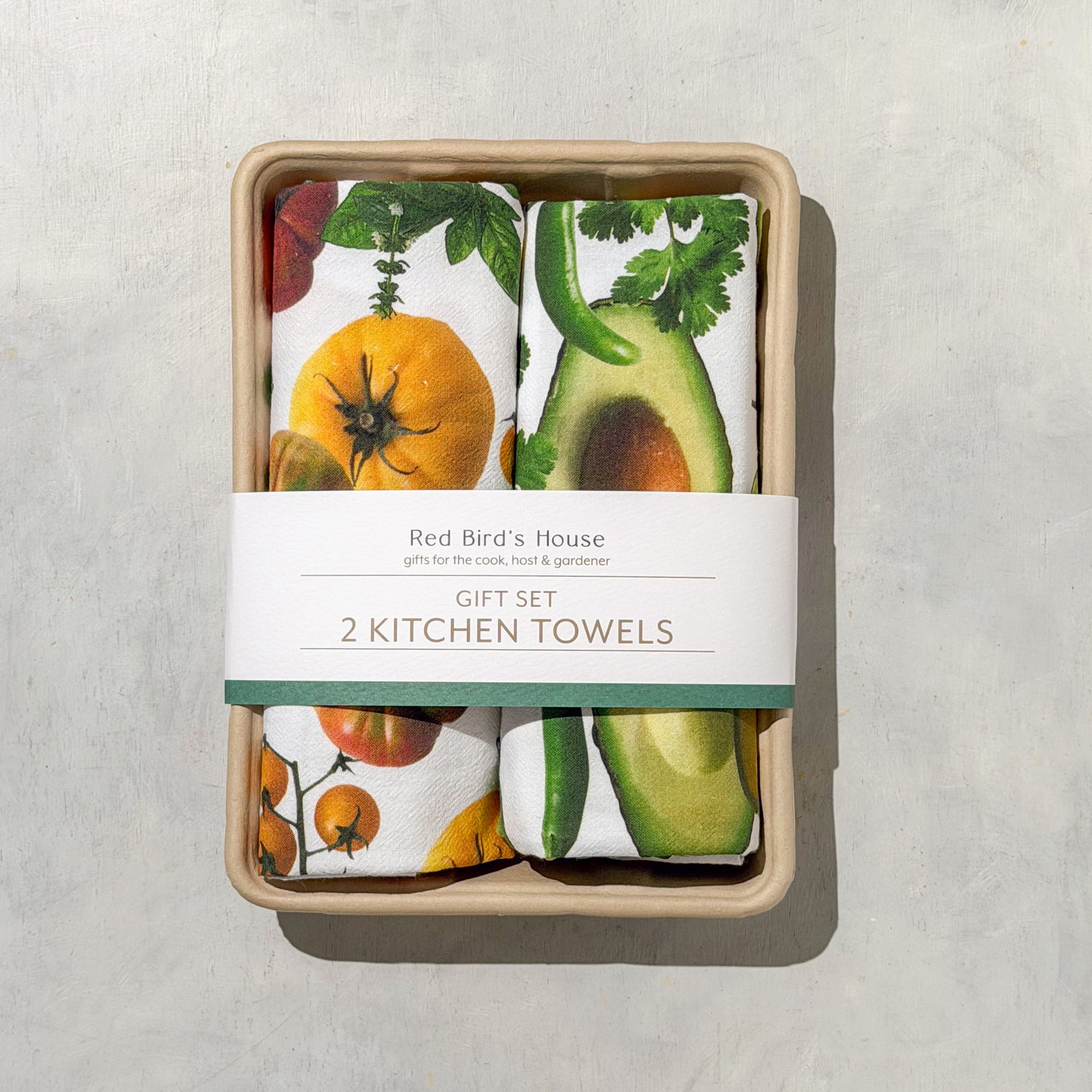 A gift set of two folded kitchen towels by Red Bird's House, packaged in a light wood tray with a white paper band label. The towels feature a colorful botanical print with tomatoes, avocado, and herbs on a white background, set against a neutral gray textured surface.