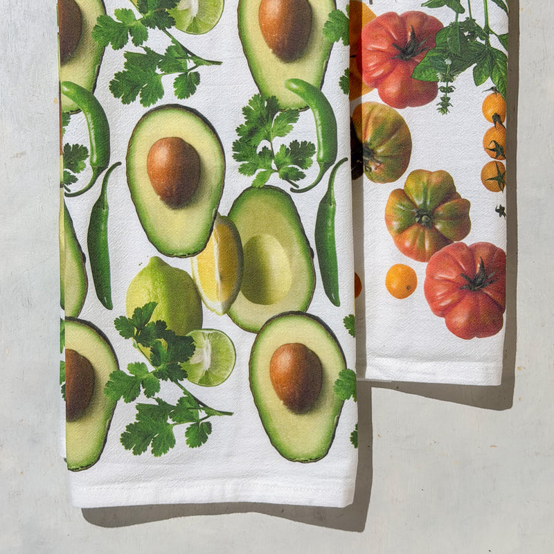 A gift set of two folded kitchen towels by Red Bird's House, packaged in a light wood tray with a white paper band label. The towels feature a colorful botanical print with tomatoes, avocado, and herbs on a white background, set against a neutral gray textured surface.